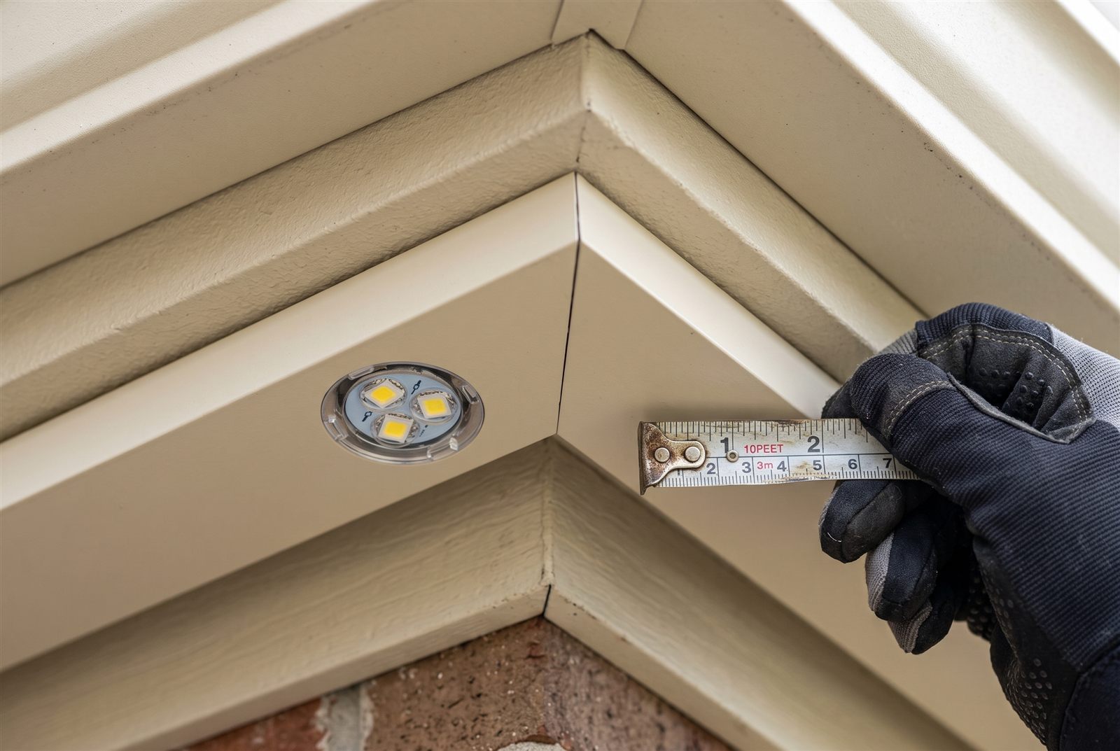 Close-up of a gloved hand holding a tape measure beside a small recessed LED light set into the cream-colored soffit trim at a roofline corner
